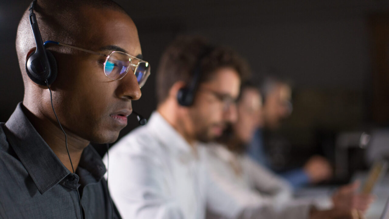 Concentrated call center operator communicating with client. African American young man in eyeglasses looking at laptop while serving client. Call center concept