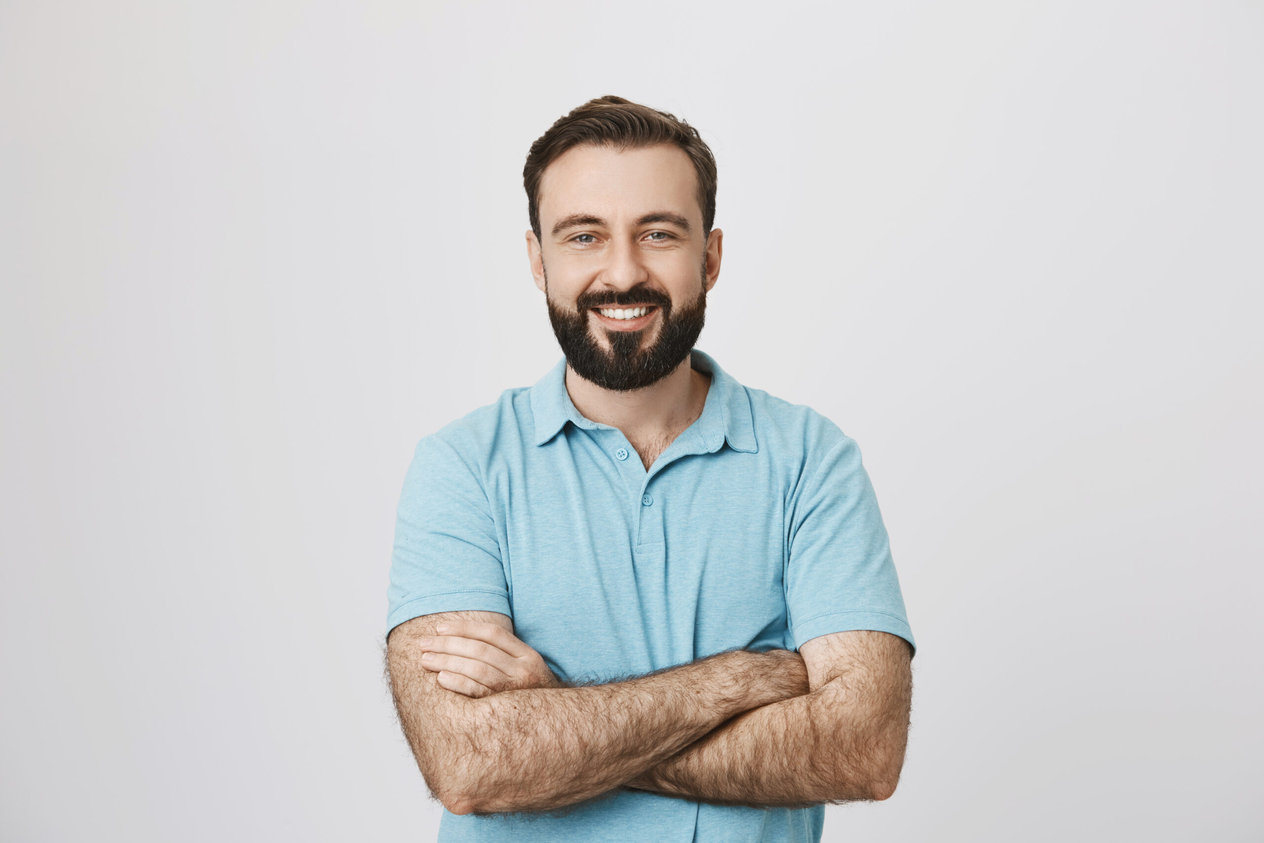 Portrait of a handsome bearded man smiling looking to the camera with his hands crossed isolated on white background. This person looks like an ordinary guy who is sincere and ready to help everyone who is in trouble.